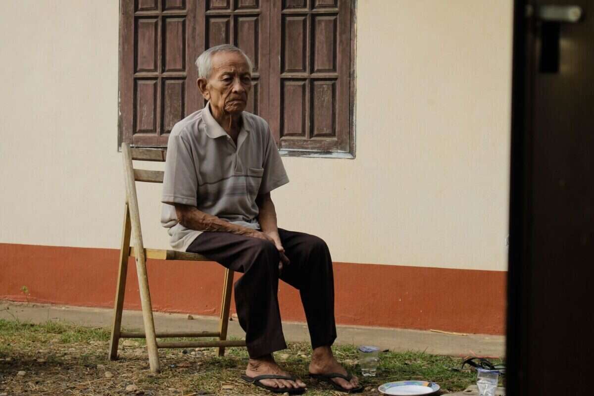 Elderly man sitting pensively on a wooden chair outside his home, reflecting solitude.